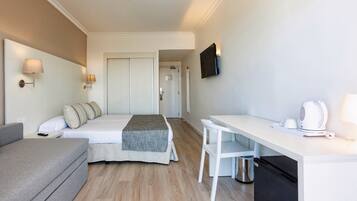 A hotel room with a bed, a sofa, a desk with a kettle, and a TV on the wall.