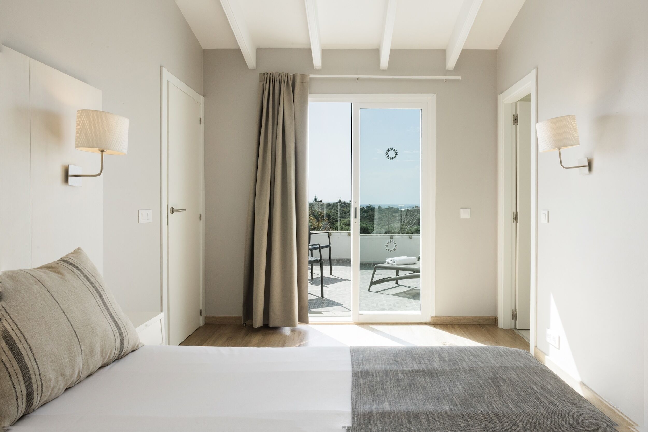 A bedroom with a bed, a window with curtains, a balcony with a table and chairs, and a clock on the wall.
