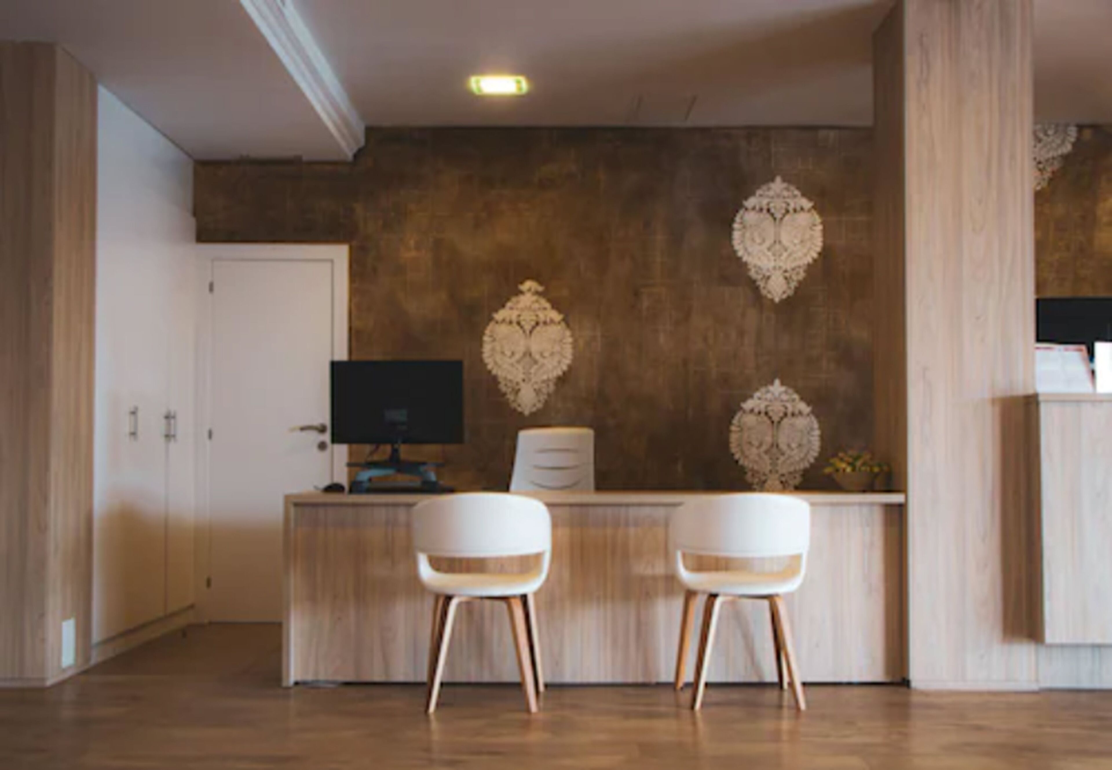 Een receptiebalie met een houten bureau, twee witte stoelen, een computermonitor en decoratieve wandkunst.