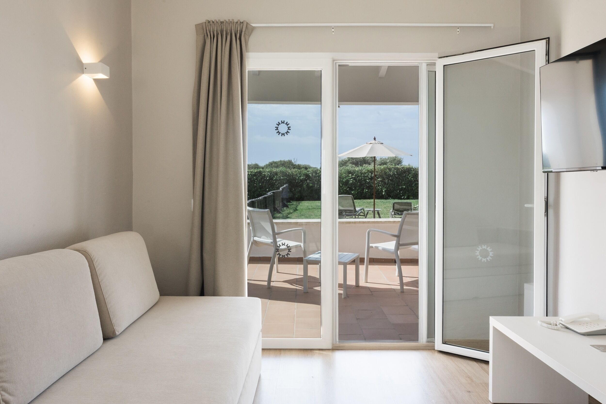 Una habitación moderna con sofá, escritorio y puerta corrediza de vidrio que da a un patio exterior con sillas y una mesa.
