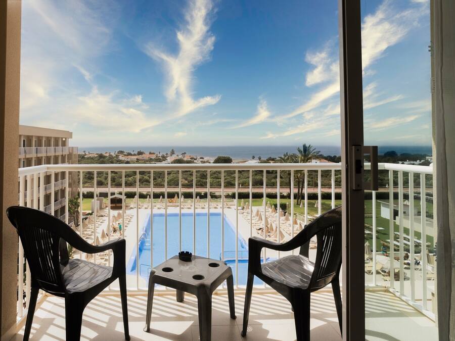 A balcony with two chairs and a small table, overlooking a swimming pool and a distant view of buildings and greenery.