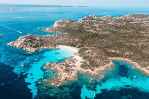 An aerial view of a coastal area with rocky terrain, clear blue waters, and a narrow sandy beach.