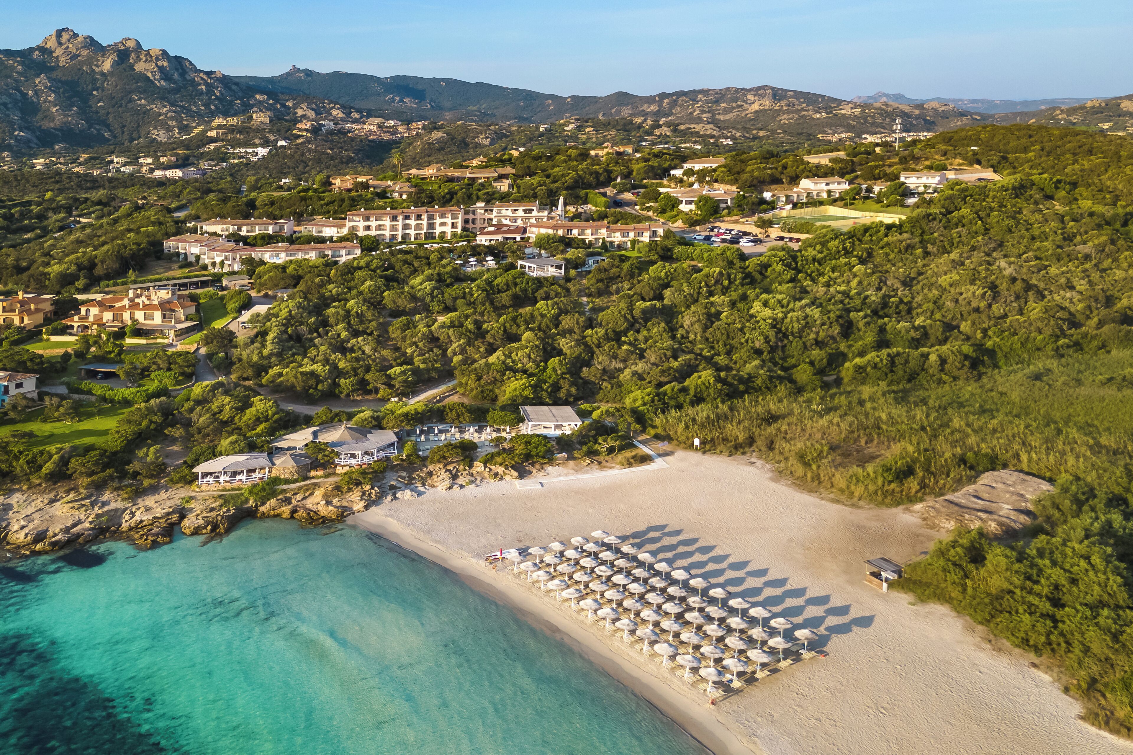 An aerial view of a coastal resort with a beach, villas, and lush greenery.