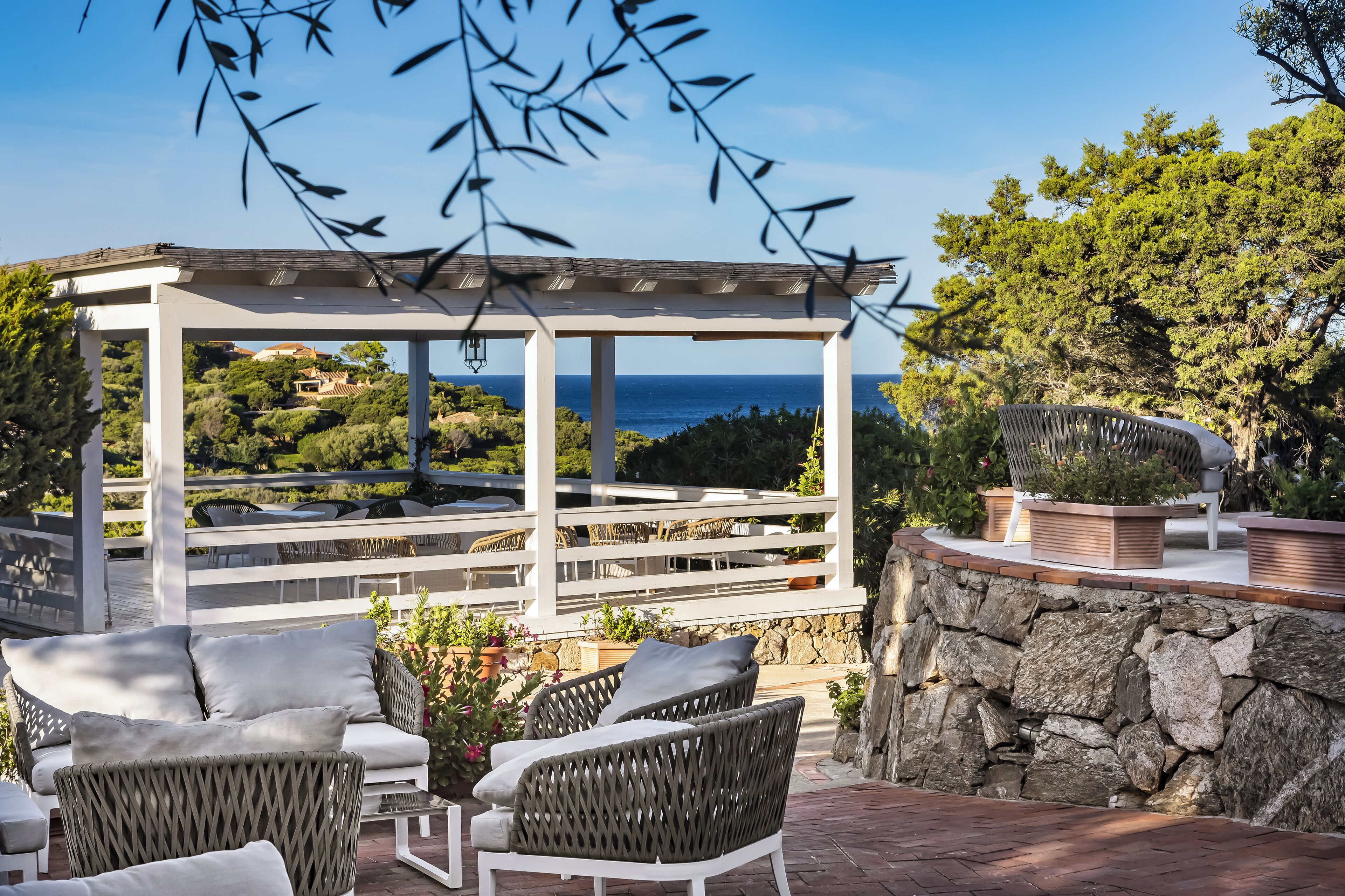 A covered outdoor seating area with wicker furniture and a stone wall, overlooking a scenic landscape with trees and the sea.