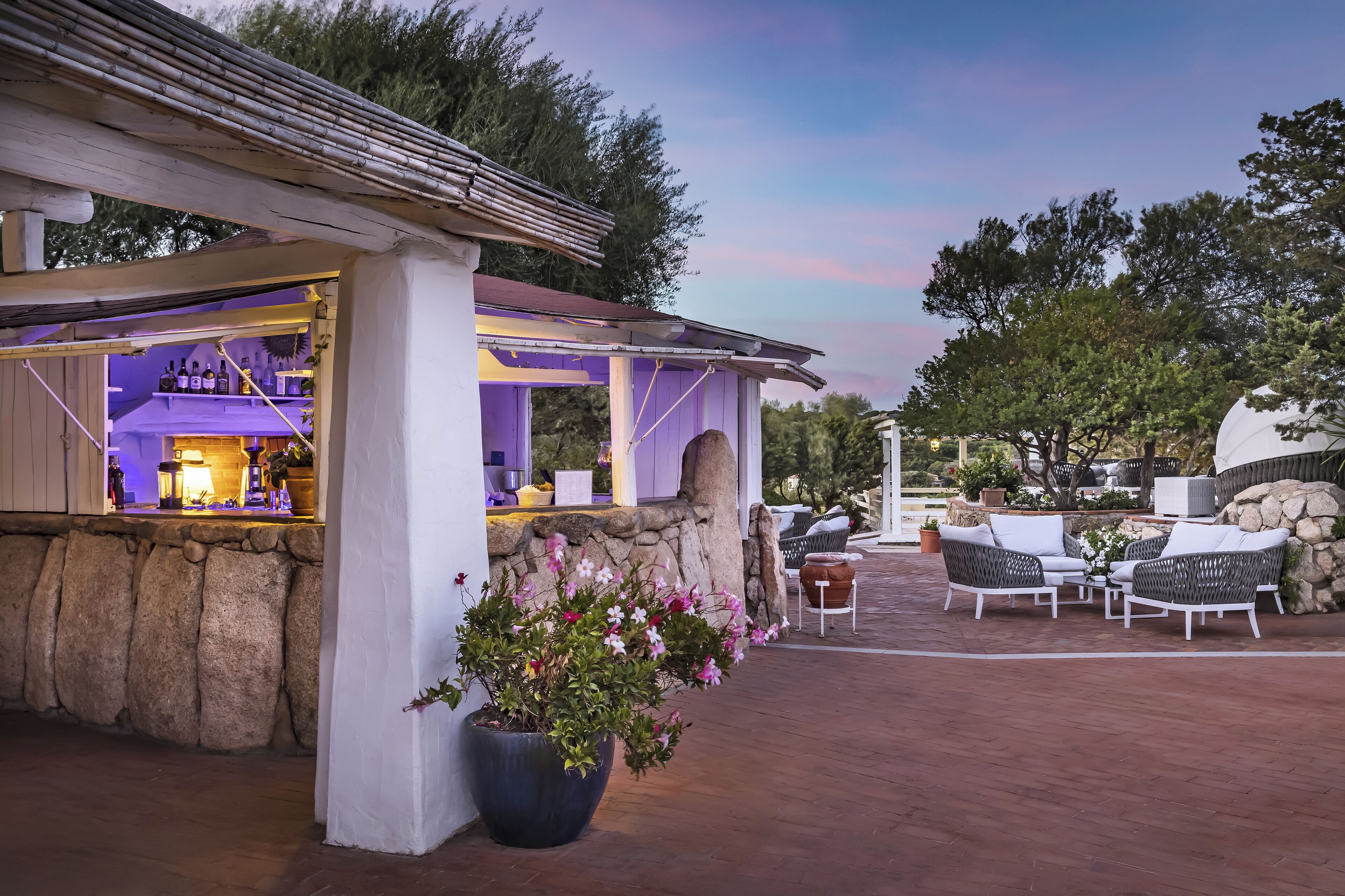 An outdoor bar area with stone walls, a large planter with flowers, and outdoor seating with white wicker furniture.