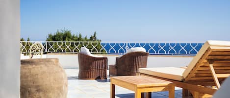 Una terraza con mobiliario de mimbre y una mesa de madera, con vista al mar azul y una valla blanca.
