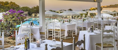 An outdoor dining area with white tables and chairs, a pool in the background, and umbrellas providing shade.