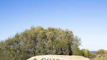 A large rock with "COSTA SMERALDA" engraved on it, surrounded by greenery and a clear sky.