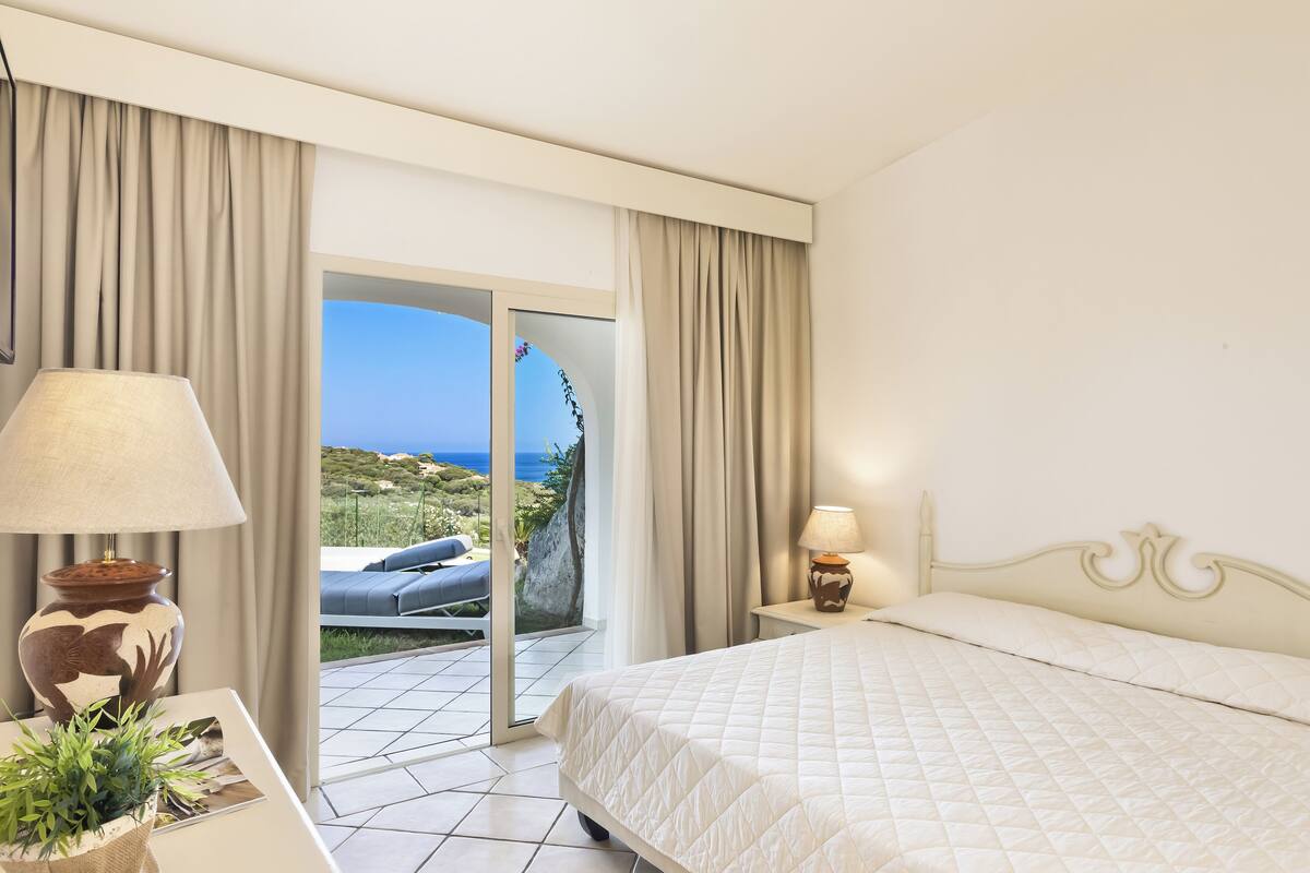 A hotel room with a bed, a nightstand, a lamp, a potted plant, and a view of the sea through a sliding glass door.