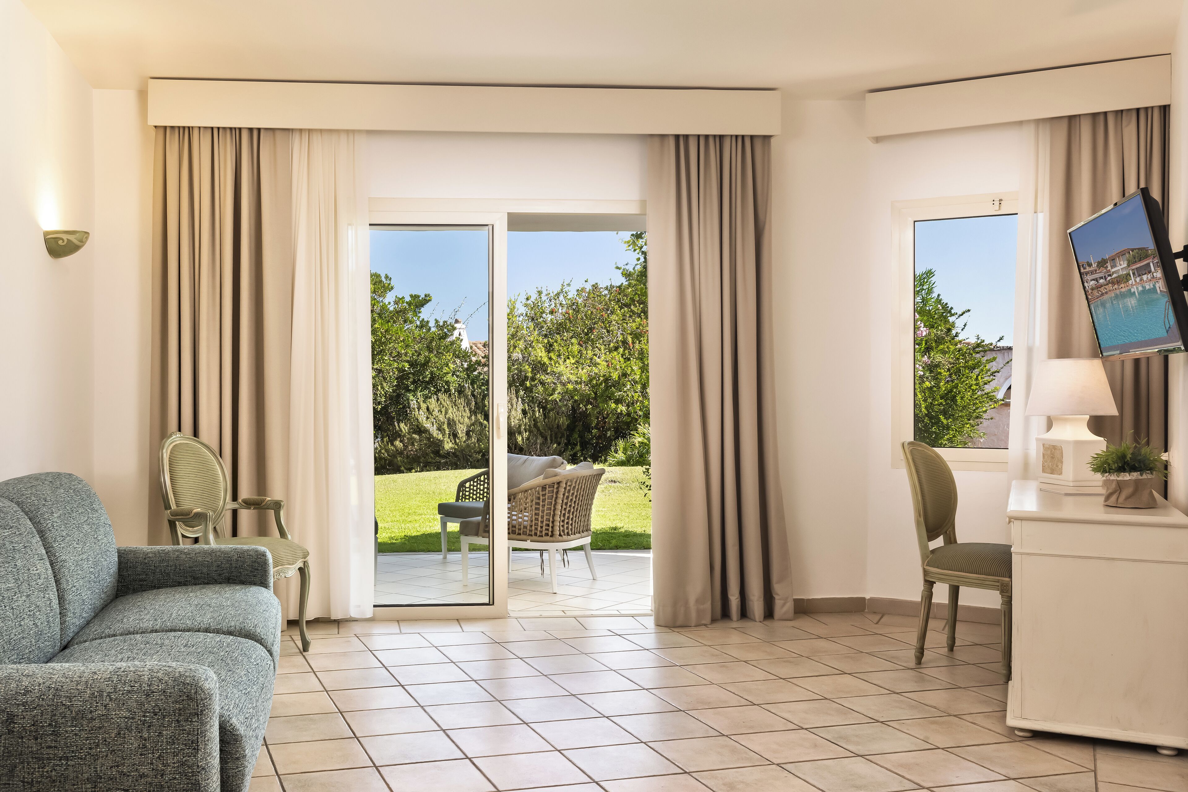 Habitación de hotel con un amplio ventanal corredero que da a un balcón, un sofá, una silla, un escritorio con lámpara y un televisor.