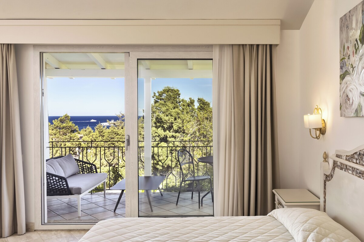 A hotel room with a bed, a nightstand, a wall-mounted lamp, and a balcony with a sofa and chairs.