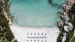 An aerial view of a beach with rows of umbrellas and lounge chairs, a small building, and a rocky coastline. - Grand Hotel in Porto Cervo (Arzachena)