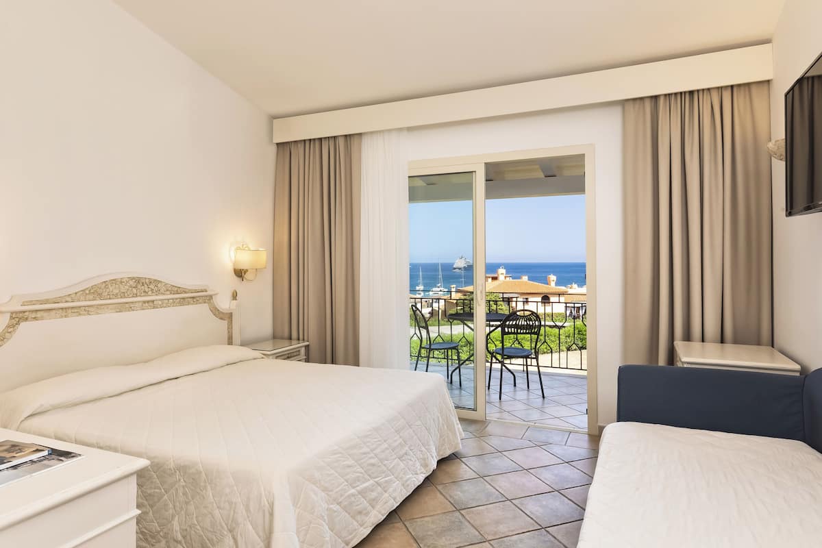A hotel room with two beds, a balcony with a view of the sea, and a dining area outside.