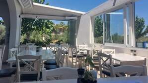 An outdoor dining area with tables and chairs, large windows, and greenery in the background. - Castro Hotel (Malevizi)