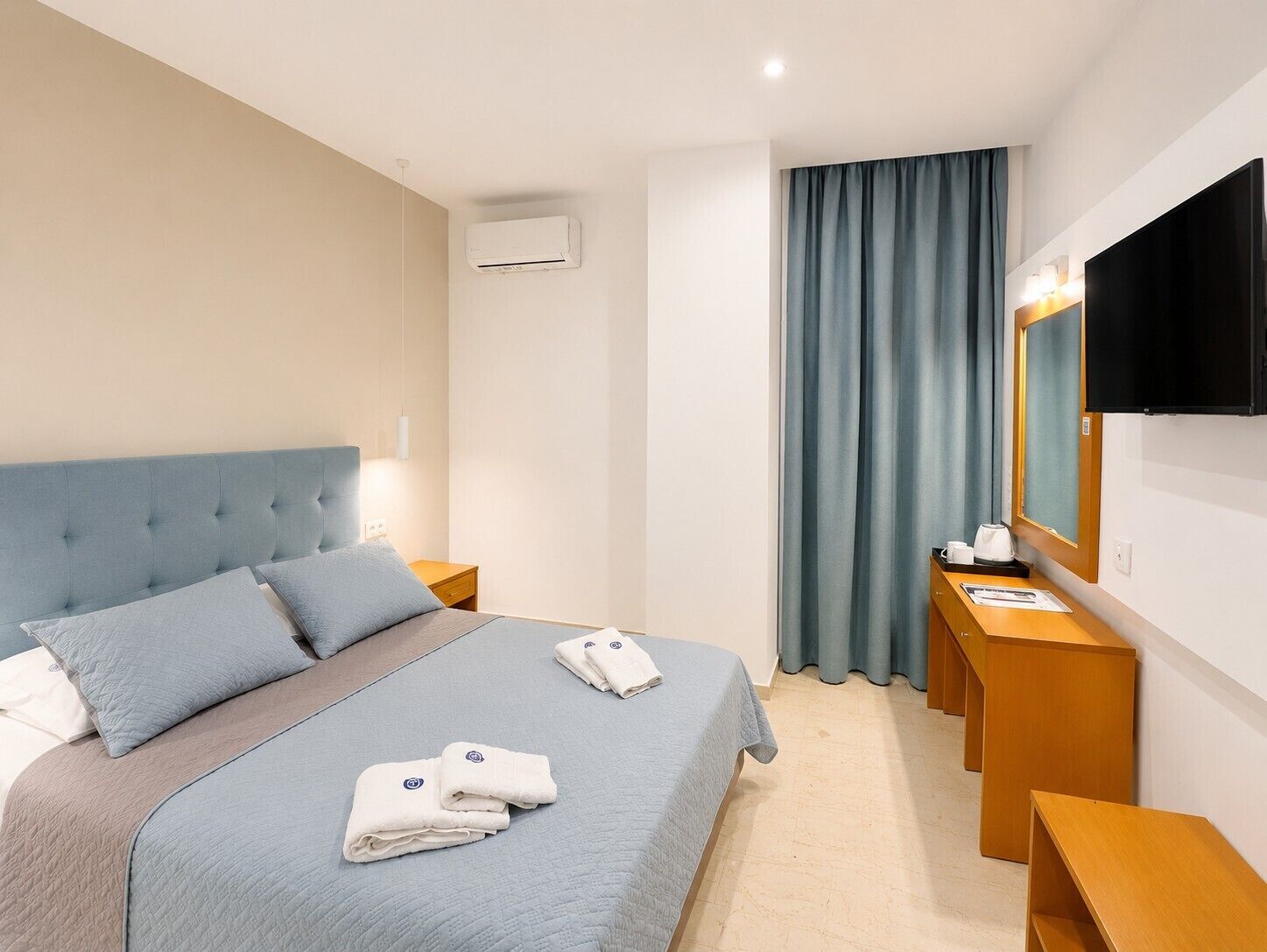 A hotel room with a bed, a bedside table, a mirror, a TV, and a window with curtains.