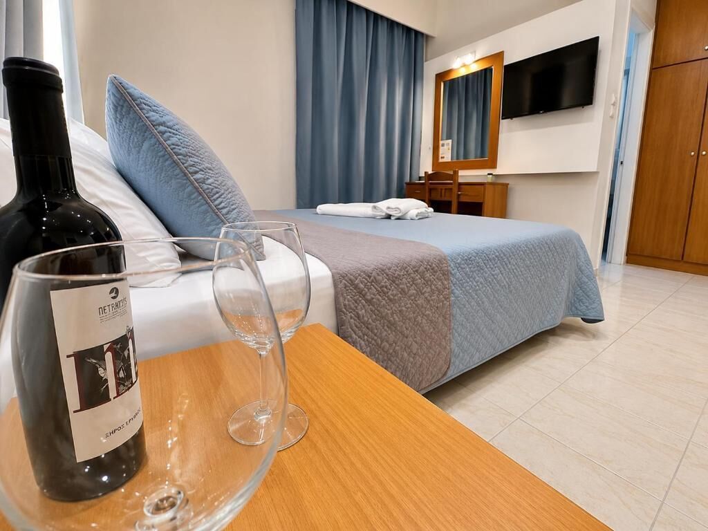 A hotel room with a bed, a wooden table with a wine bottle and glasses, a TV, and a wardrobe.