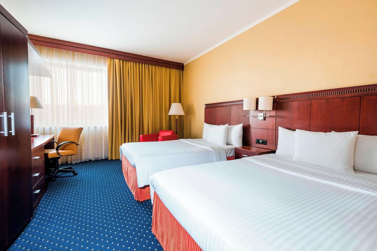 Ein Hotelzimmer mit zwei Betten, einem Schreibtisch, einem Stuhl und einem großen Fenster mit Vorhängen.