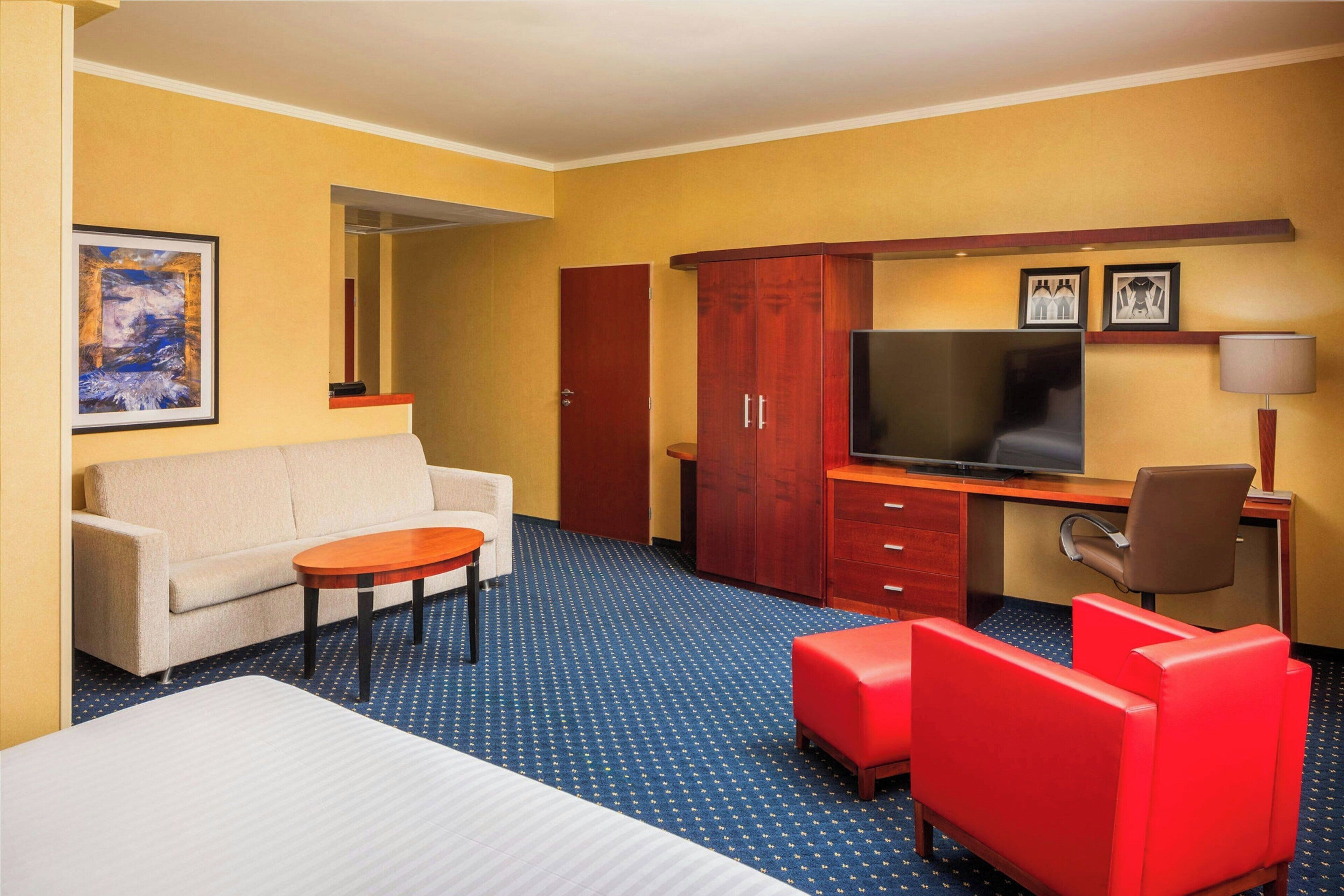 A hotel room with a bed, a sofa, a desk with a TV, a red chair, and a wooden wardrobe.