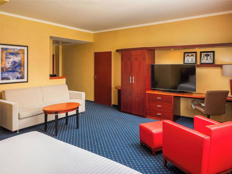 A hotel room with a bed, a sofa, a desk with a TV, a red chair, and a wooden wardrobe.