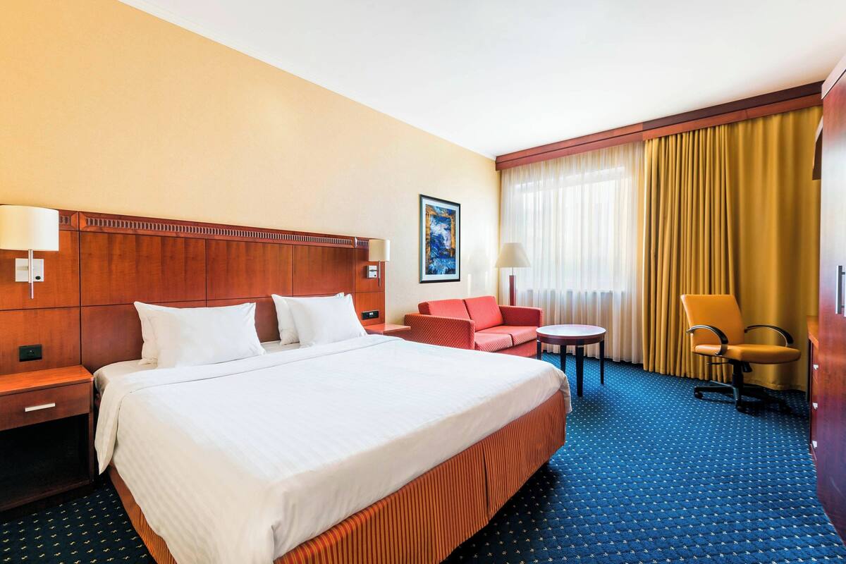 Ein Hotelzimmer mit einem großen Bett, einem hölzernen Kopfteil, einem roten Sofa, einem kleinen runden Tisch und einem gelben Vorhang.