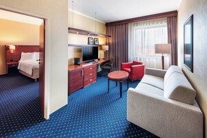 Quarto de hotel com uma cama, uma escrivaninha com TV, uma cadeira vermelha, um sofá cinzento e uma janela grande com cortinas.