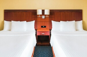 Quarto de hotel com duas camas, cabeceira de madeira, mesa de cabeceira com duas luminárias e uma pequena gaveta.
