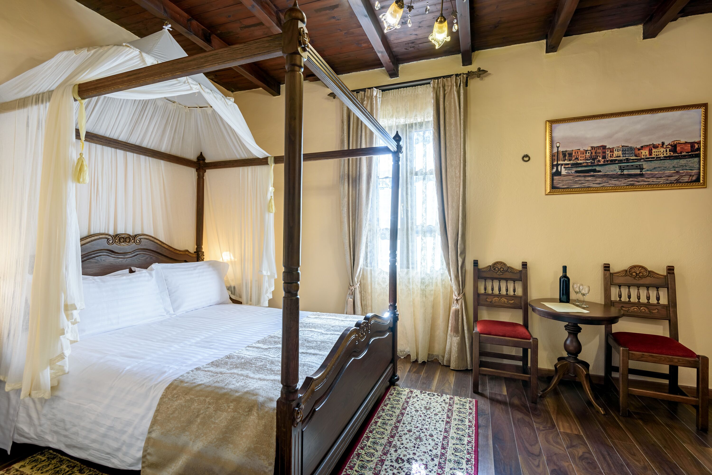 A four-poster bed with a canopy, a wooden chair with a red cushion, a small round table, and a painting on the wall.