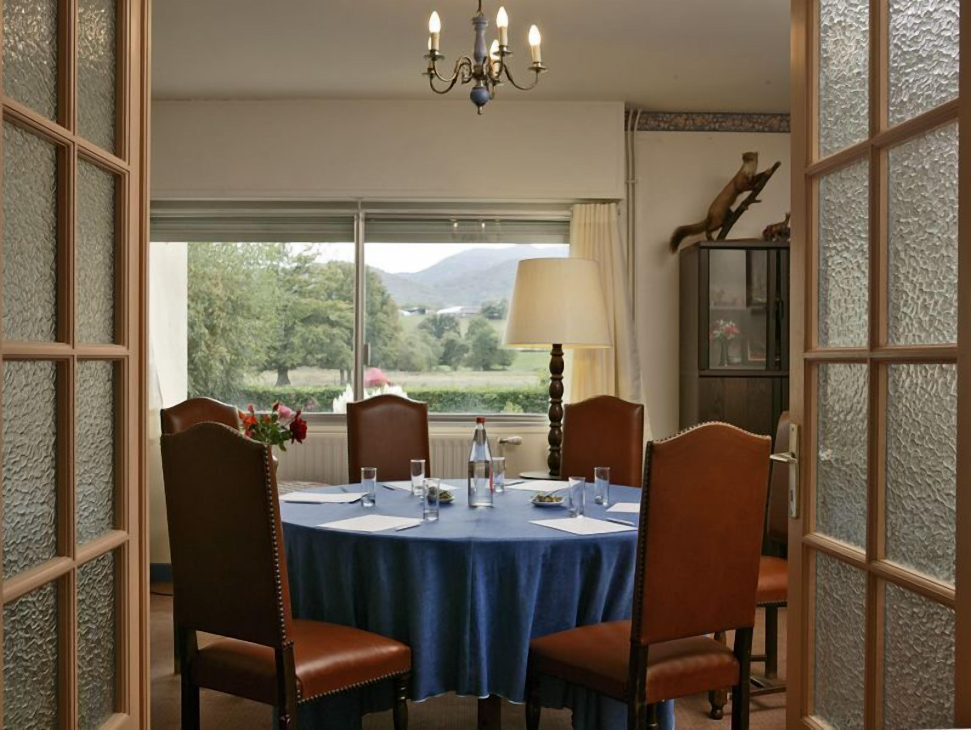 A dining area with a round table, four chairs, a chandelier, a window with a view of greenery and mountains, and a lamp.