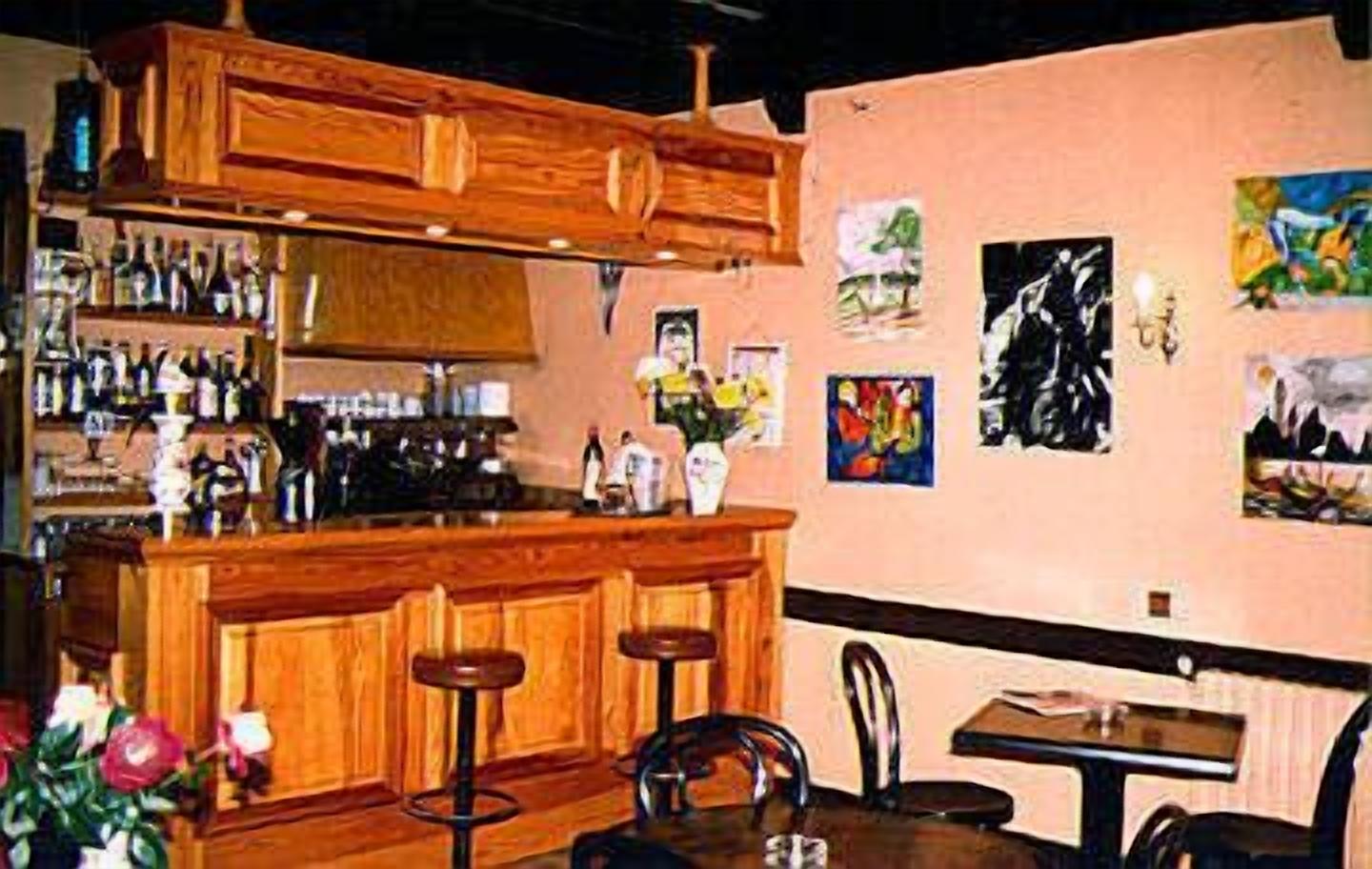Un área de bar con armarios de madera, una barra y taburetes. Hay pinturas en la pared y una mesa con sillas.