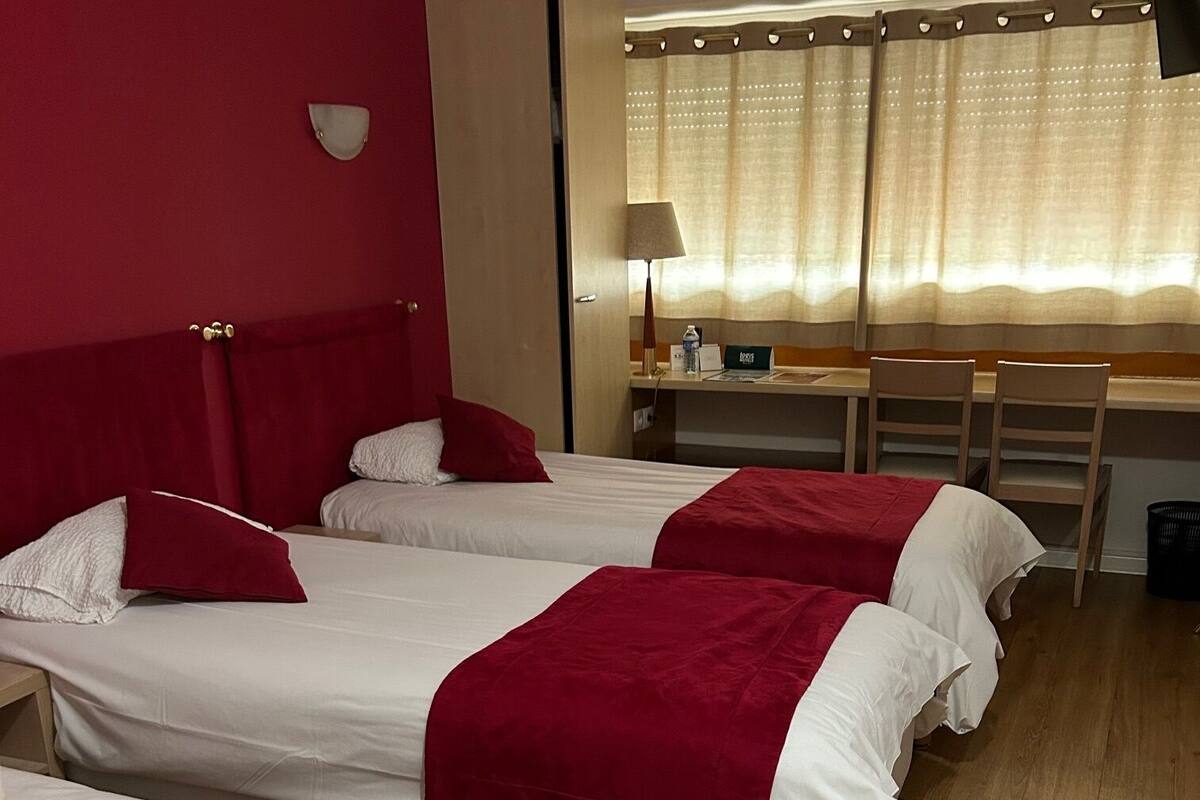 A hotel room with three single beds, a desk with a chair, a lamp, and a window with curtains.