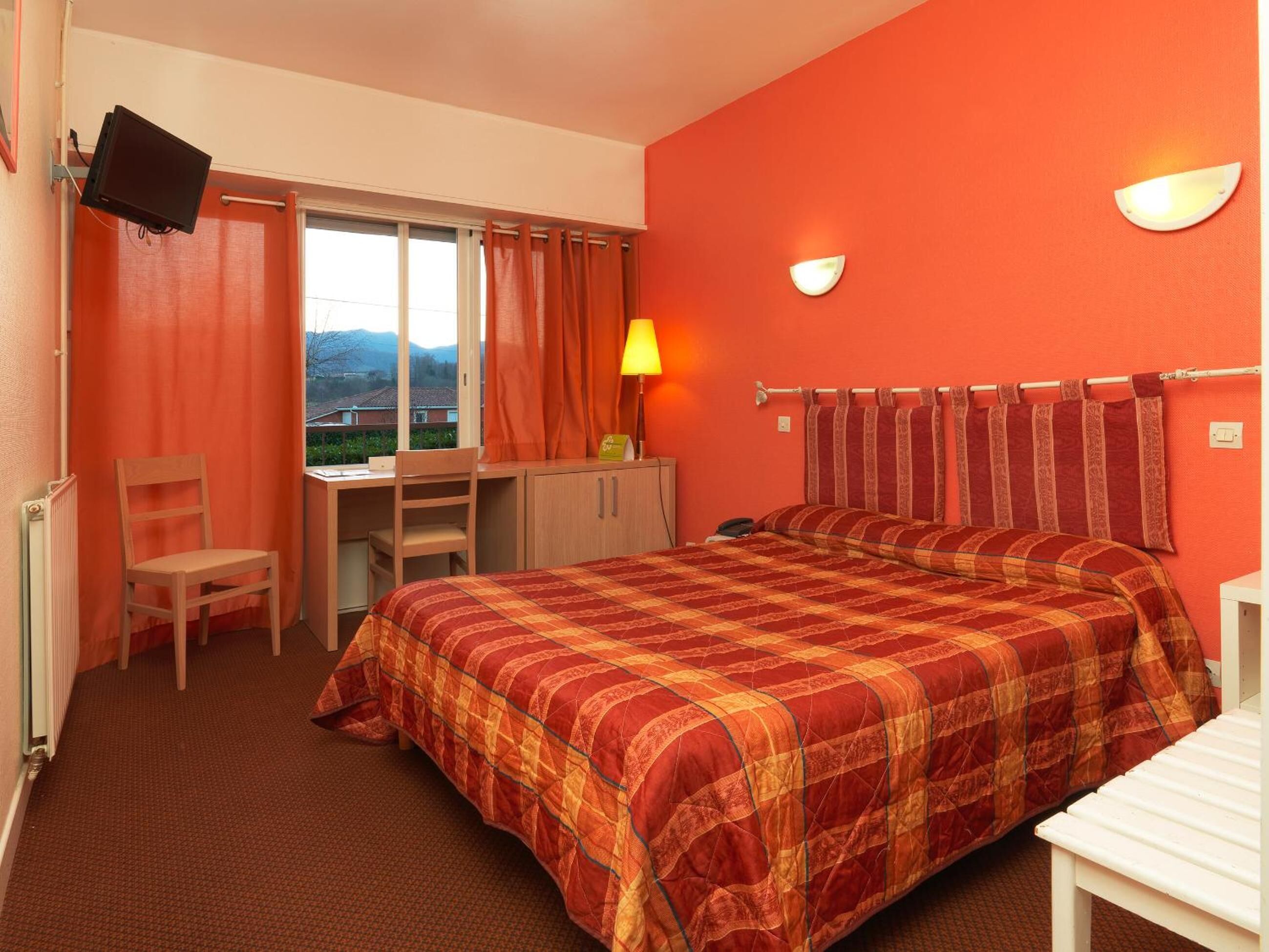 Una camera d'albergo con un letto, una scrivania, una sedia, una TV e una finestra con tende.