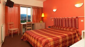 Una camera d'albergo con un letto, una scrivania, una sedia, una TV e una finestra con tende.