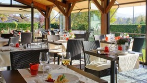 Een terras met tafels en stoelen, een houten plafond en uitzicht op groen.