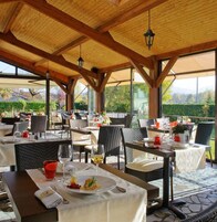 Un área de comedor al aire libre con mesas y sillas, techo de madera y vistas a la vegetación.