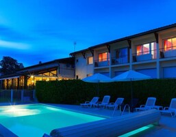 Una piscina con sillas de descanso y sombrillas, un edificio con balcones y ventanas, y un seto al frente.
