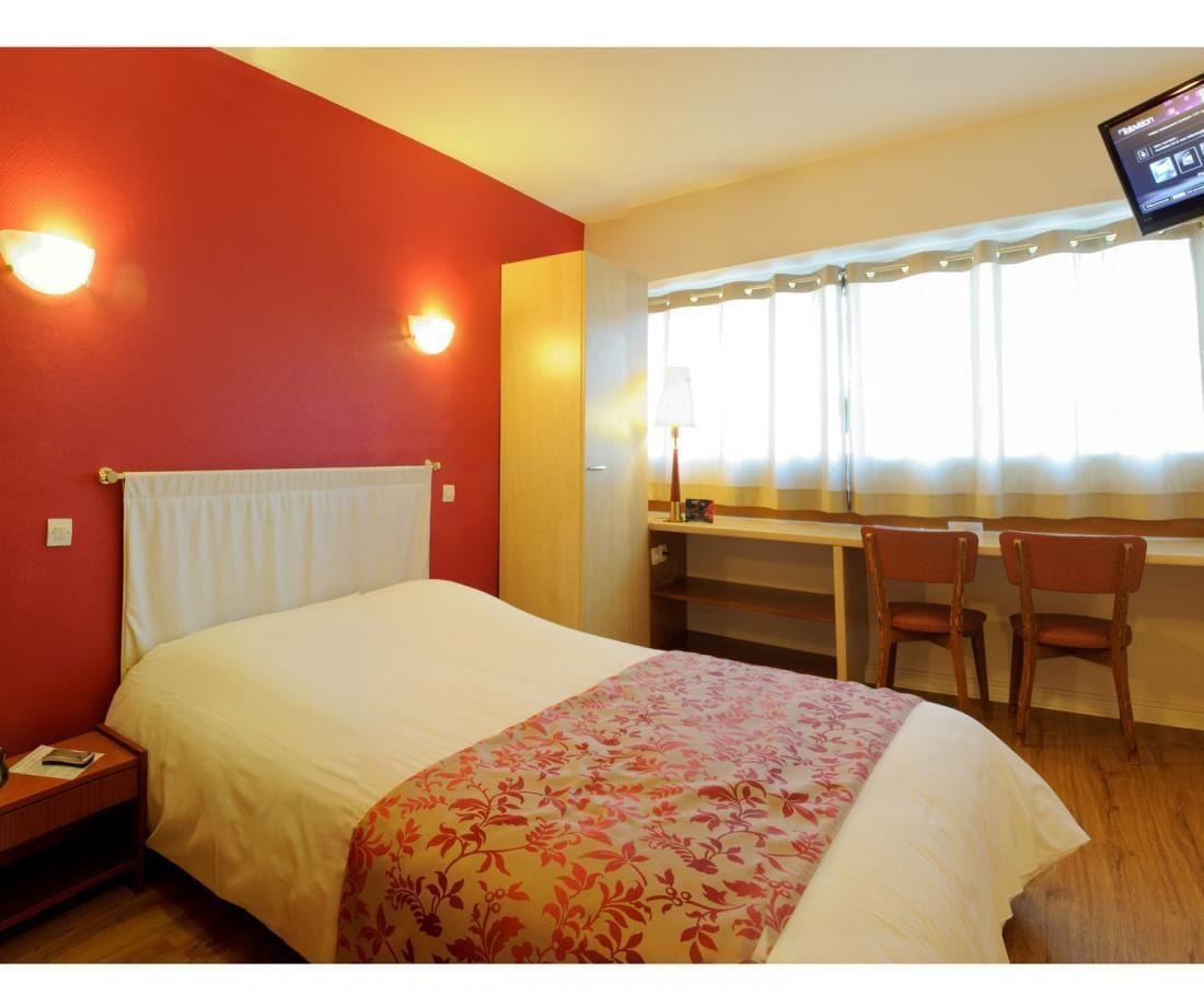 Quarto de hotel com uma cama, uma escrivaninha, duas cadeiras, uma televisão e uma janela com cortinas.