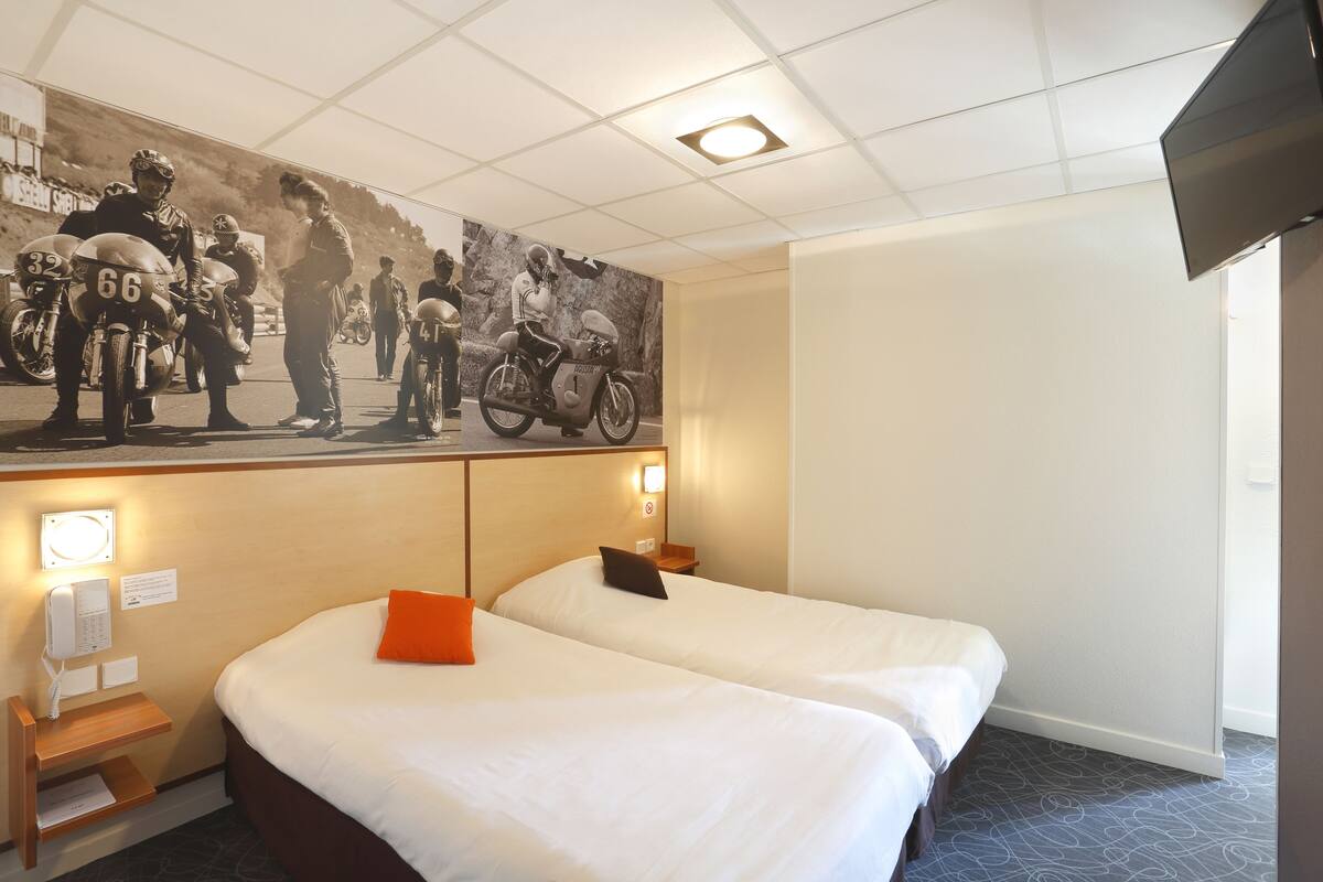 A hotel room with two beds, a mural of motorcycle racers, a flat-screen TV, and a telephone.