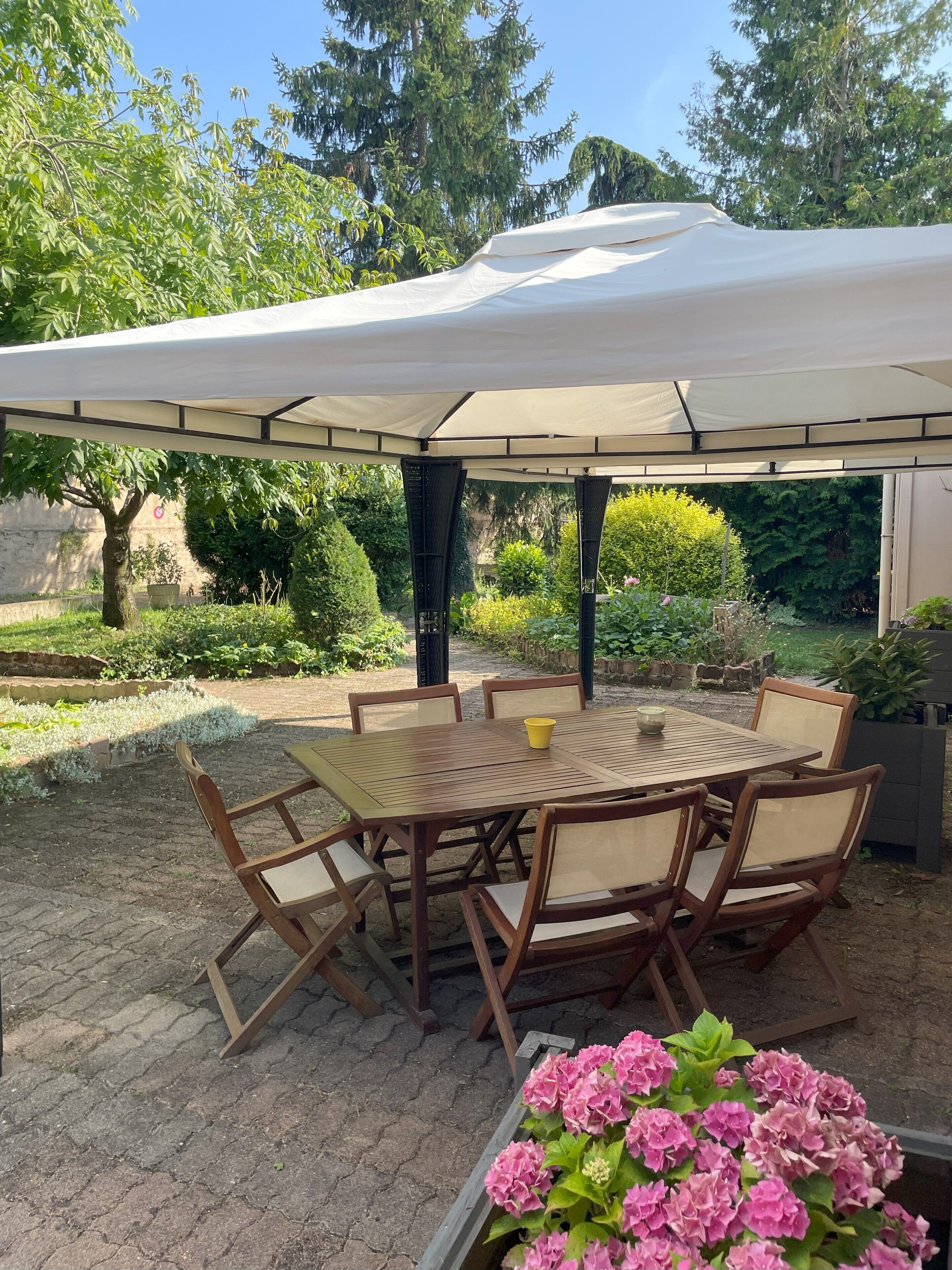 An outdoor patio with a round wooden table and six chairs, covered by a large white canopy.