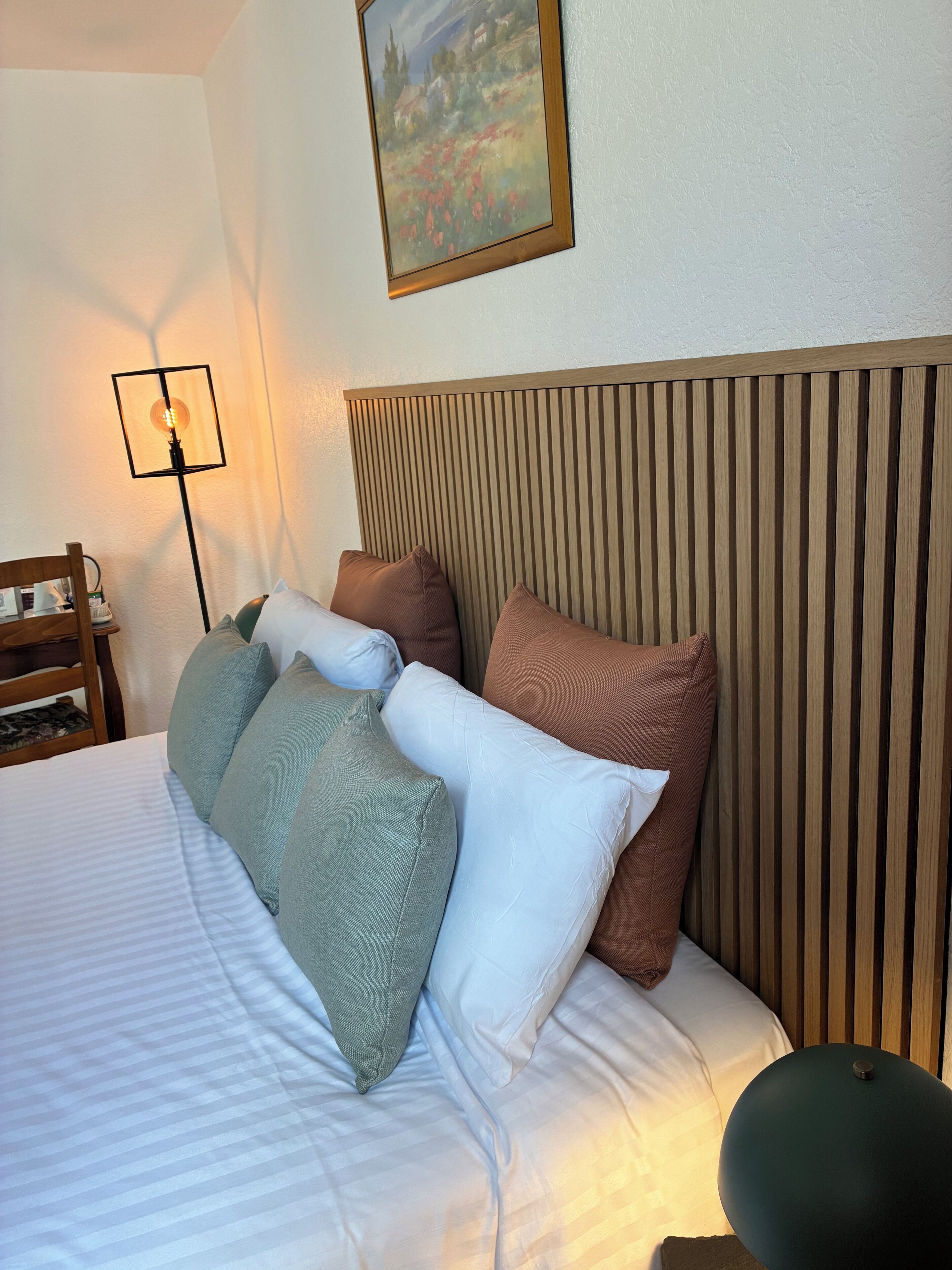 Un lit bien fait, avec des oreillers, une table de chevet sur laquelle se trouve une lampe, et un tableau accroché au mur.