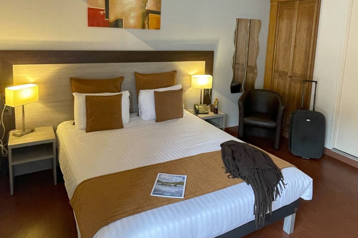 Habitación de hotel con una cama grande, dos lámparas de noche, un cabecero de madera, un cuadro abstracto colgado en la pared, un armario de madera, una silla negra y una maleta.