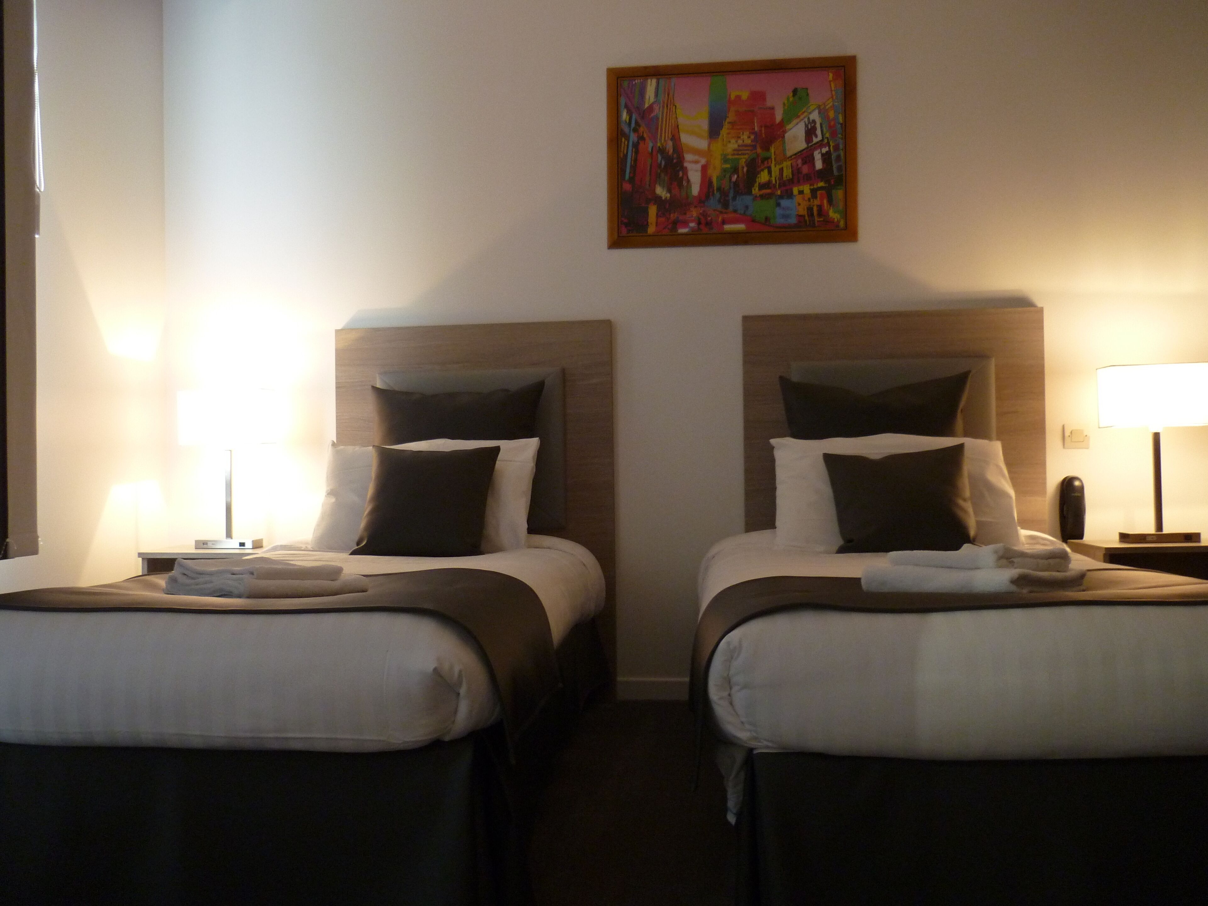 A hotel room with two beds, each with a wooden headboard, white bedding, and a single bedside table with a lamp.