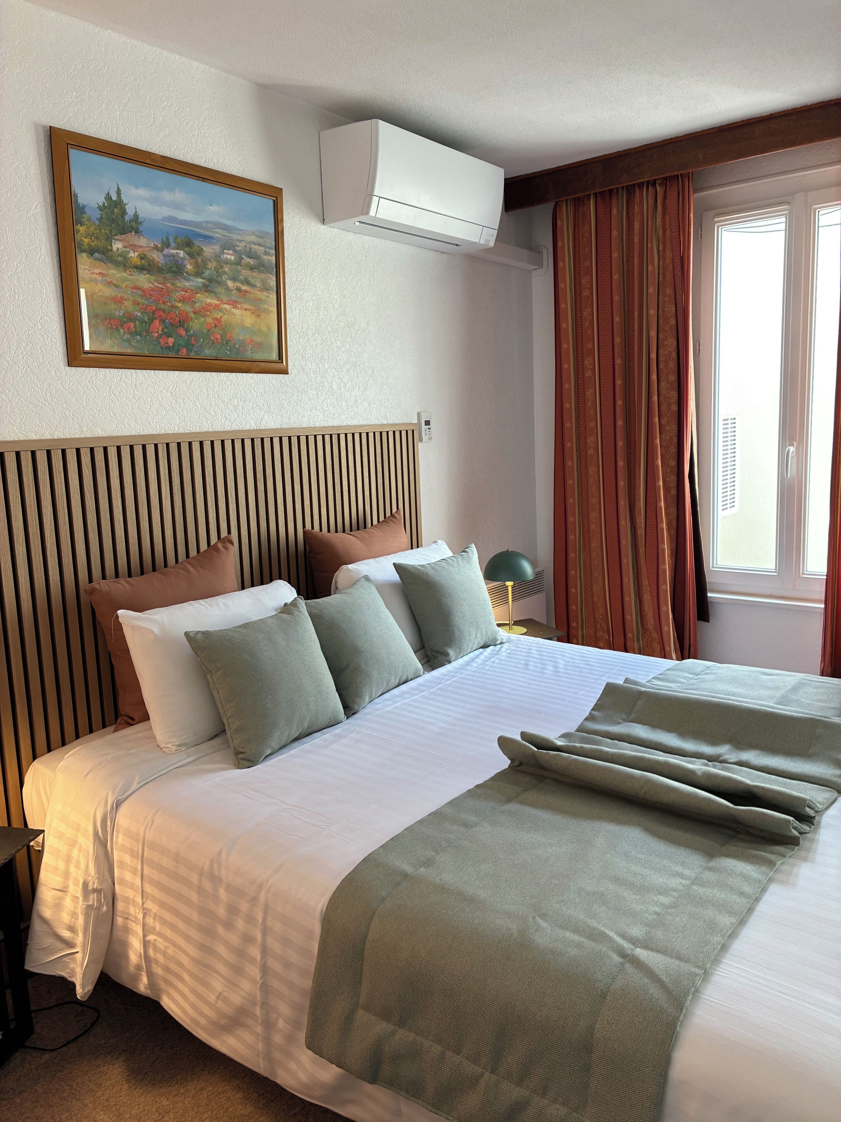 Una habitación de hotel con una cama, un cuadro, aire acondicionado, una ventana con cortinas y una lámpara de noche.