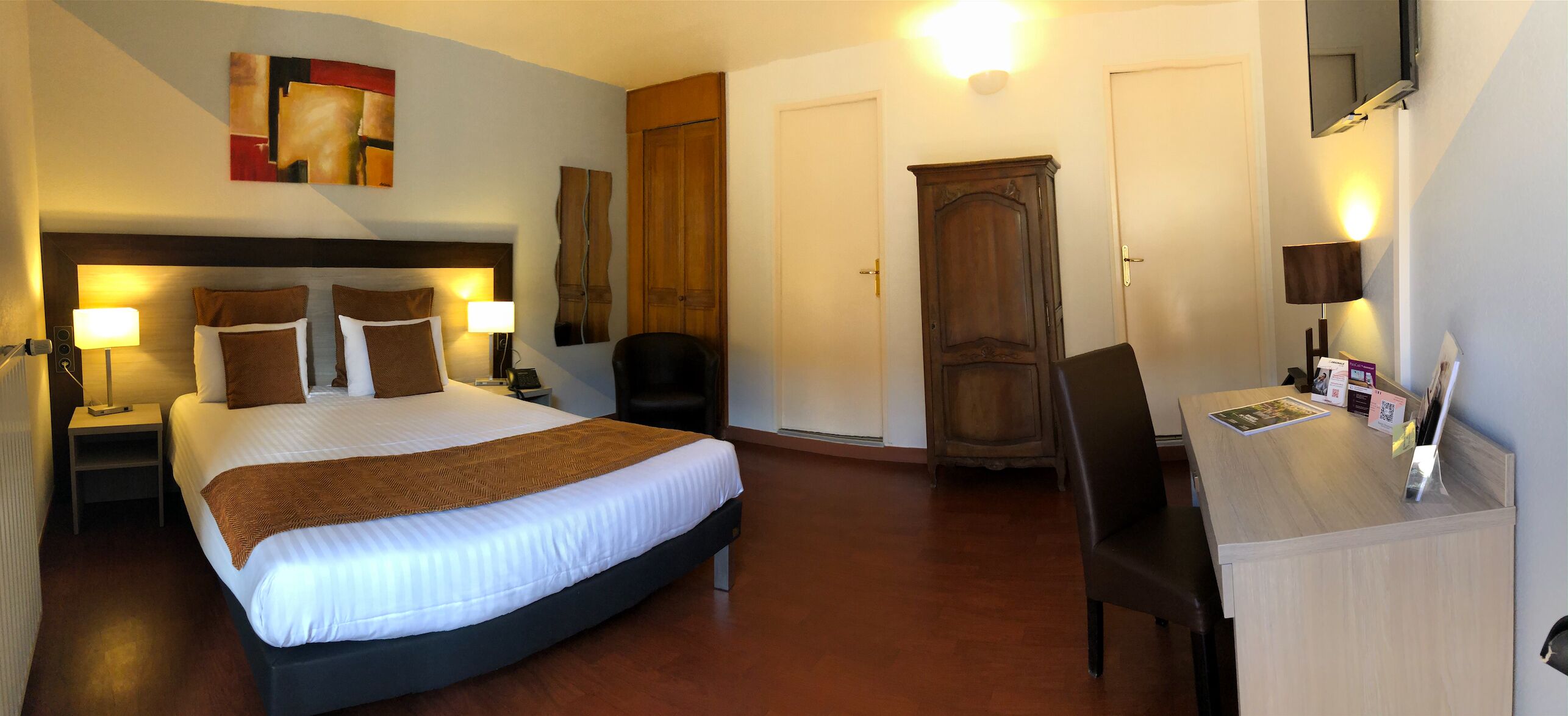 Quarto de hotel com uma cama grande, duas mesas de cabeceira com luminárias, um guarda-roupa de madeira, uma escrivaninha com cadeira e um espelho.