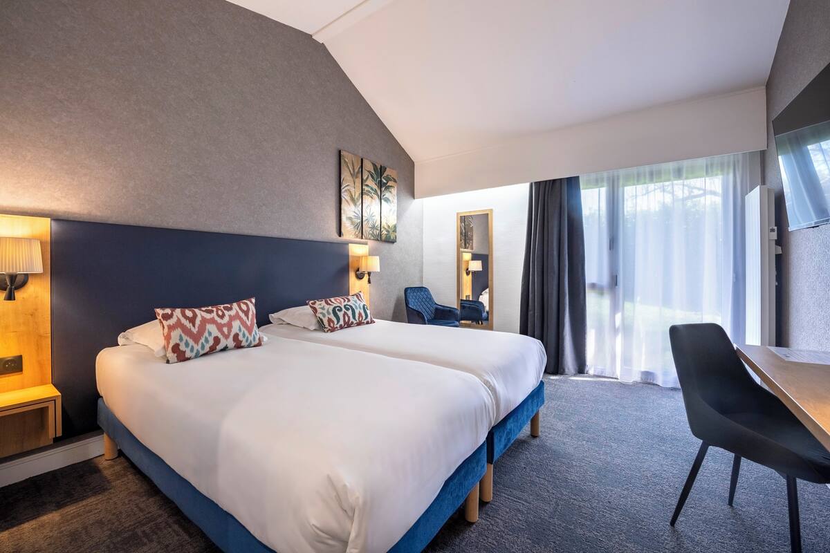 Une chambre d’hôtel avec deux lits, un bureau, une chaise et une grande fenêtre avec des rideaux.