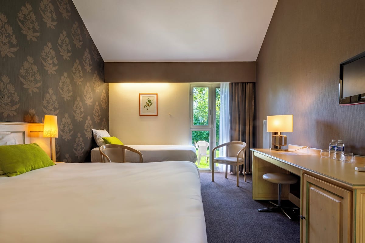 Une chambre d’hôtel avec deux lits, un bureau, une chaise, une télévision et une fenêtre donnant sur un espace verdoyant.