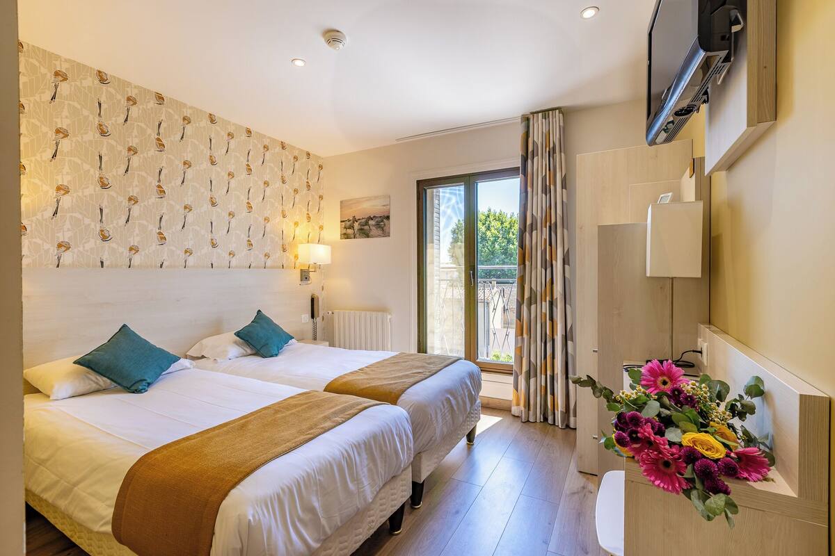 A hotel room with two beds, a large window with curtains, a wall-mounted TV, and a flower arrangement on a side table.
