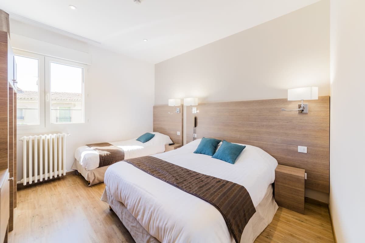 A hotel room with a large bed, a smaller bed, two bedside lamps, a wooden headboard, and a window with a view of buildings.