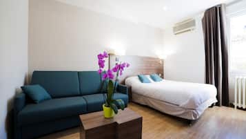 Una habitación de hotel moderna con sofá, cama, una mesita de madera con una planta y una ventana con cortinas.