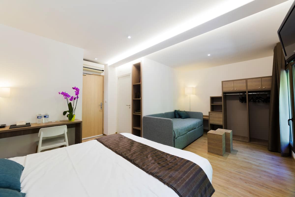 A modern hotel room with a bed, desk, chair, sofa, wardrobe, and a small table with a vase of flowers.