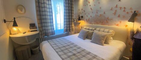 Un dormitorio con cama, mesita de noche, lámpara, ventana con cortinas, pared decorada con siluetas de pájaros y radiador.